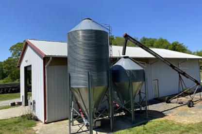 A white metal farm building stands beside two galvanized grain silos of different sizes. An auger conveyor extends from the ground to the top of the smaller silo, while trees and a clear blue sky form the background. Gravel and patches of grass surround the agricultural structures.